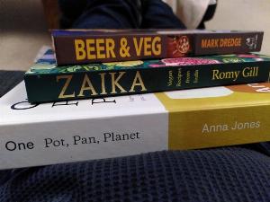 The spines of three books visible with the torso and legs (both clothes in dark blue shades) in the background.  The books are "Beer & Veg" by Mark Dredge,  "Zaika: Vegan recipes from India" by Romy Gill and "One Pot, Pan, Planet" by Anna Jones.