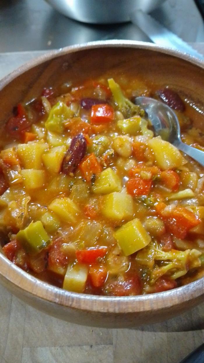 A red hued stew with numerous vegetables and legumes in a bamboo bowl with a stainless steel spoon