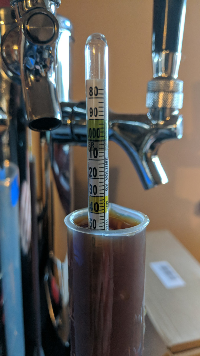 A close up of a hydrometer measuring gravity of a brown beer with some chrome bar taps in the background