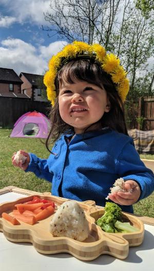 A 2 year old girl, wearing a blue cardigan and dandelion crown, stands in front of a plate with carrots, broccoli and onigiri - eating onigiri