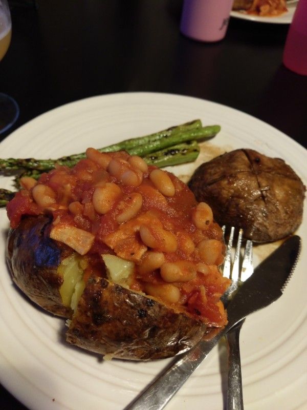 A large baked potato with crispy skin, loaded with homemade baked beans.  Behind sits a glazed Scottish flat mushroom and some grilled asparagus spears.