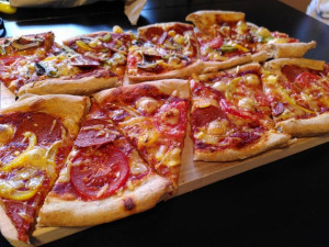 長方形の竹製まな板の上に、2枚のピザから16枚のスライスを載せています。 前列はシンプルなヴィーガン・ペパロニに3種類の'チーズ'、後列は同じだがハラペーニョとマッシュルームが入っている。