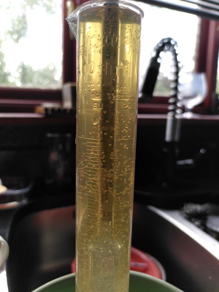 A plastic measuring vessel containing a very clear pale ale in the foreground.  in the background is a brown wooden window sill, a double window with blurred view and a large kitchen sink tap surrounded by coiled chrome.