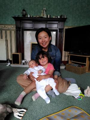 A Japanese woman with short hair wearing a denim jacket, kneeling with a three year old Scottish and Japanese girl who is holding a new born Japanese baby.