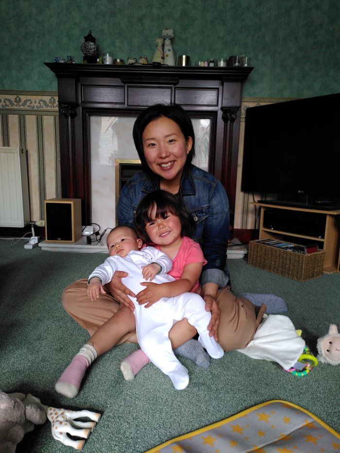 A Japanese woman with short hair wearing a denim jacket, kneeling with a three year old Scottish and Japanese girl who is holding a new born Japanese baby.
