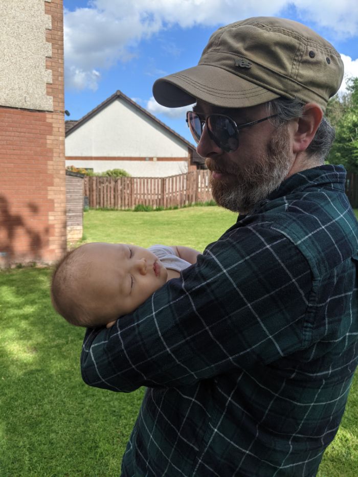 A bearded guy, wearing a checked blue and green shirt, beige cap and sunglasses cradling a three monrh old baby in his arms. it's a sunny day with a residential garden view in the background.
