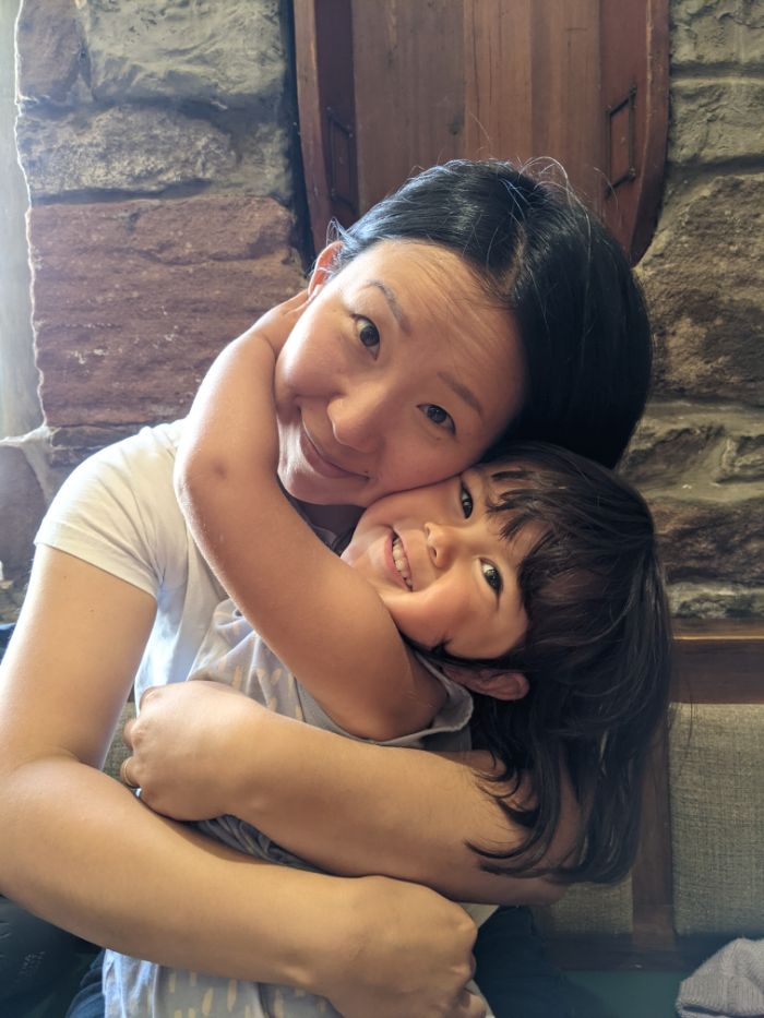 An Asian woman and her young daughter hugging in, in the background is a bare stone wall, the interior of a bar.