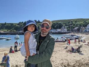 A middle-aged white guy with gingery white beard, wearing a black t-shirt, green jacket, jeans, sunglasses and a cap carrying his young daughter in his arms, she is wearing a blue summer dress and blue hat.  the background is a small but fairly crowded sandy beach with some boats in the water.