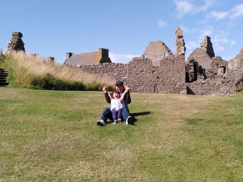 An Asian woman sits on a grassy hill with her daughter between her legs, hands clasped and reaching for the sky.  in the background are some ancient castle ruins.