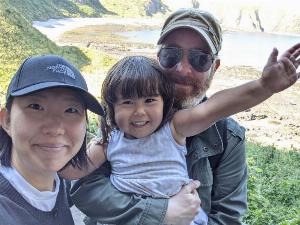 a selfie, with an Asian woman wearing a black cap, a white guy with a gingery white beard wearing a beige cap and sunglasses carrying their daughter, wearing a blue Summer dress with shoulder length brown hair, between them. in the background and at a much lower elevation is a rocky beach cove.