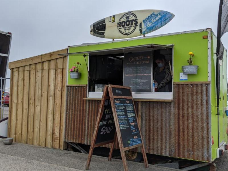 A largely wooden food truck with green upper half, a surfboard sits atop the truck with the text "Roots - Plant-based catering" and "Roots @ The Beach".  There is a menu A-board in front of the truck, some flowers in aluminium pails, two kiosk windows,one for orders and one for collection and a maske woman is visible inside the truck.  In the foreground is the promenade path.