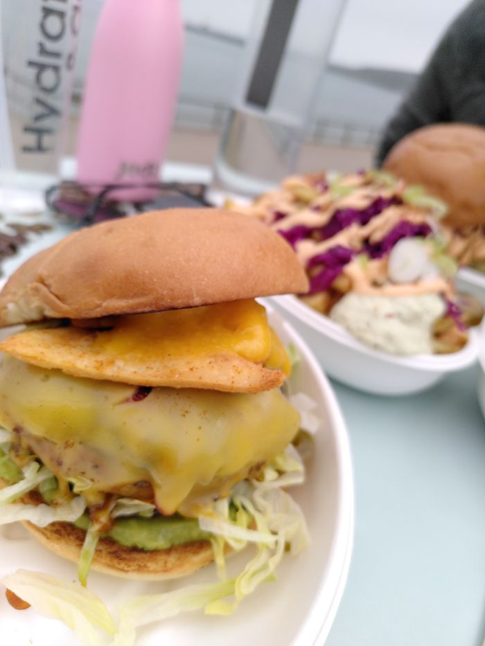 The foreground is a shot of a Plant-based burger, with vegan cheese and some nachos atop of the patty and lettuce & avocado below,  in the background is a bowl of loaded fries covered with sriracha mayo, pickled cabbage, a white sauce and spring onions. further back is a blurry burger, detailed in the next shot.