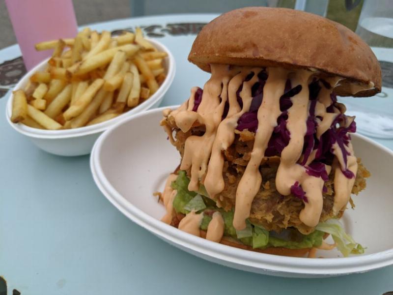 The foreground is a shot of a Plant-based burger, with pickled red cabbage atop of the patty and lettuce & avocado below, a mess of light orange sriracha mayo runs down the sides of the burger.  in the background is a bowl of fries.