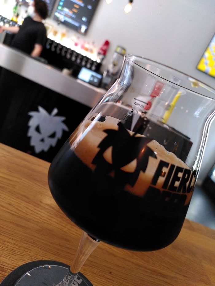 A tilted shot of a Fierce Beer branded glass with a dark beer inside, the bar is visible in the background with a steel Fierce Hopman logo,  some blurred taps, a menu monitor and a masked barman with curly dark hair.