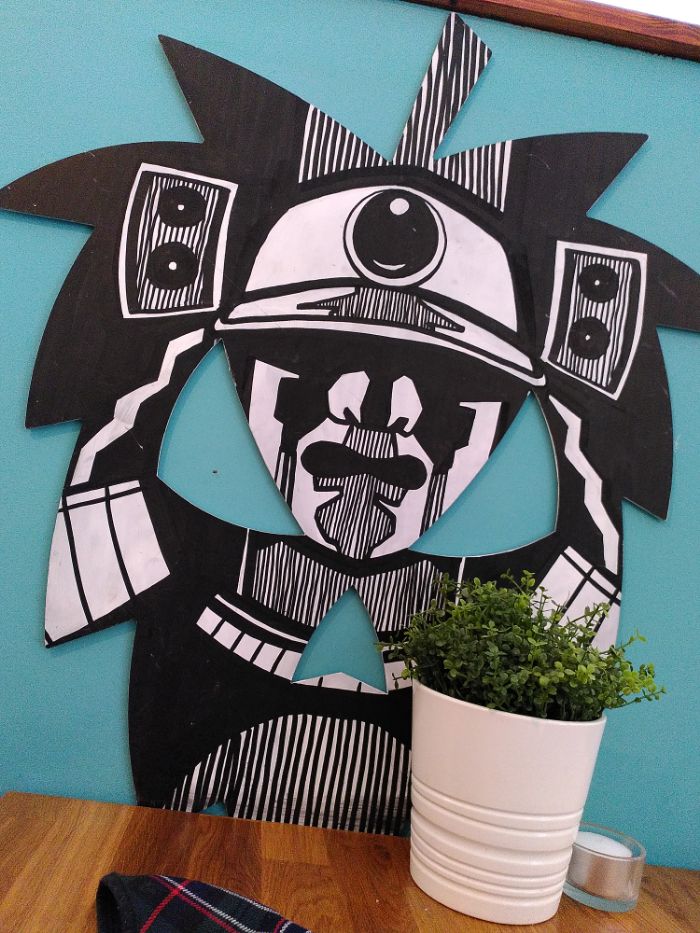 A samurai-themed decorated cutout of the Fierce Hopman logo on a blue wall.  A small, herbaceous looking plant in a white pot sits in front of the logo, next to a small candle.  A bit of a black watch tartan face mask is also visible on a wooden table.