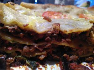 A slightly mis-focussed side on shot of a lasagne, showing three layers of pasta, green lentils in a tomato and aubergine sauce, a layer of spinach, a layer of baby courgette from our garden, a cashew bechamel-esaque sauce and some slice tomato on top.