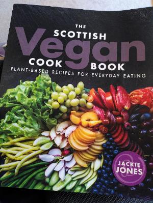 The front cover of The Scottish Vegan Cookbook by Jacki Jones, top half of cover is black with the book title in white and grey text.  the lower half of the cover is a very colourful array of prepared fruit, veg and leafy greens, dark red cherries and purple blueberries  to light green broadband and lettuce leaves.
