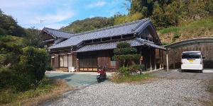 A ground level shot of a traditional Japanese wooden house.  There is a nicely manicures tree in front of the house and next to it a red moped.  To the right of the house under the car shelter is a white family car.  An orange or red mailbox can be seen on a pillar at the corner of the house.  There are several tiers of old-fashioned black roof tiles.