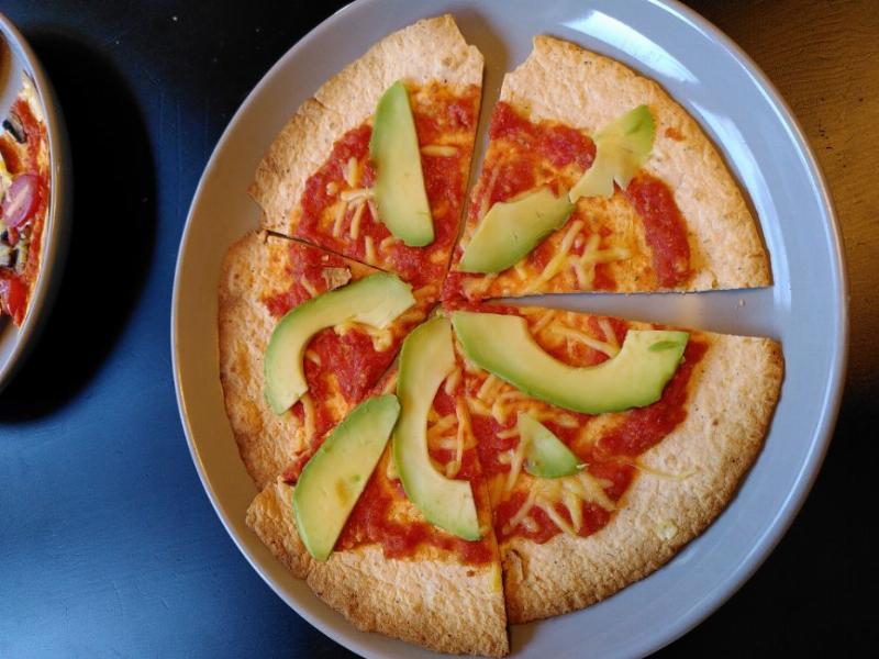 a tortilla based pizza with tomato sauce and thin slices of avocado. it sits on a grey plate atop a black wooden table.