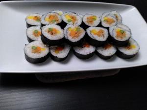 A white plate on a black table holding many maki sushi, the fillings are carrot, yellow pepper, asparagus and ginger soy mince.  