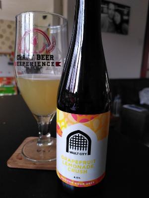 a brown beer bottle with white, yellow, orange and red label.  The logo is silhouette of an old fashioned vault gate, the brewery is Vault City. The beer is a 4.0% modern sour beer called grapefruit lemonade crush.  slightly behind the bottle to the left is a beer glass with the words craft beer experience and some red graphics printed on it, inside is a slightly opaque lemon yellow beer. the background is a blurred living room scene. 
