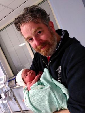 A tired, somewhat greying middle aged man white a white and ginger beard, wearing a black hoodie, stands proudly holding his newborn baby daughter.  She is wrapped in green blankets and has a pink and white wooden hat upon her perfect wee head.