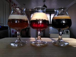 Three stemmed (goblet style) glass schooners with the word BEER printed on them in black letters. In the left is a slightly opaque, orange hewed pale ale with a white head.  The middle glass holds a roasted tea coloured brown ale with large creamy white head. The last glass holds a black stout with light brownish bubbly head.  the glasses are on a light beige bamboo chopping board. the background is a blurred kitchen scene.