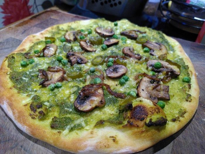 A lightly baked, floury based, thin hand-stretched pizza on a brown chopping board.  The toppings are green wild garlic pesto, brown chestnut mushroom slices, slightly caramelised onions, green peas, sliced jalapeños and a sprinkling of vegan grated cheese.