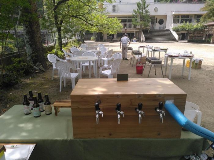 The rear of a temporary bar stall, including a wooden box enclosing a pourer with 4 shiny taps.  Ahead is a BBQ area currently devoid of customers but with space for 15 including 3 BBQs.  We are set up in the playground of a former school opposite the brewery.