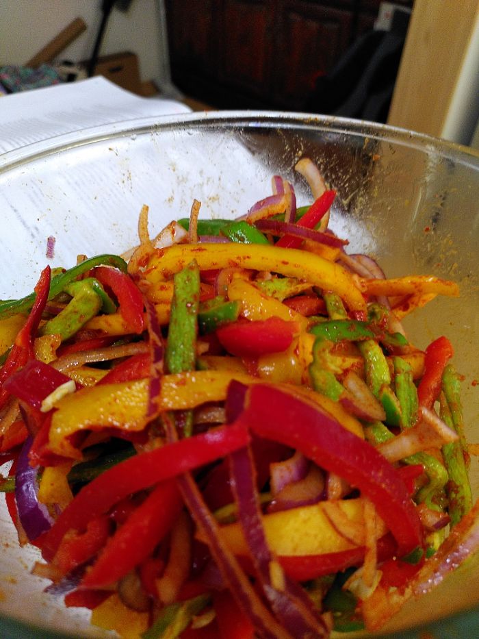 A glass bowl filled with slices of red, yellow and green peppers as well as sliced red onion all coated in reddish spices.