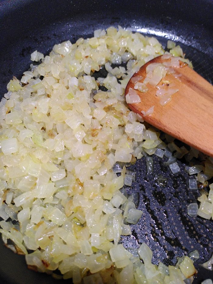 a frying pan containing chopped onions sauteed to gently browning, also a wooden implement is in the pan.