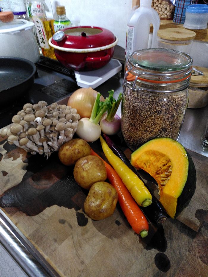 a selection of seasonal, colourful root vegetables and a jar of unpolished barley sits atop a brown wooden chopping board 