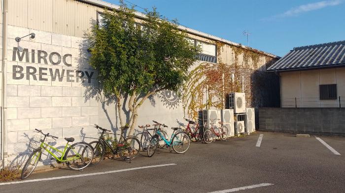 Outer wall of an industrial building with some bikes and foliage. The Miroc Beer logo is visible.