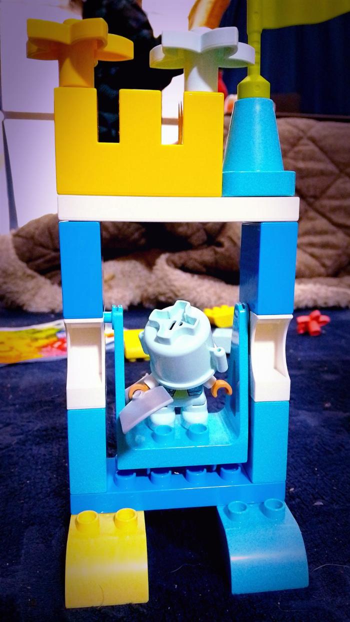 a primarily blue and yellow castle-ish swing structure with a Duplo person wearing a bucket over their head and wielding a shovel.