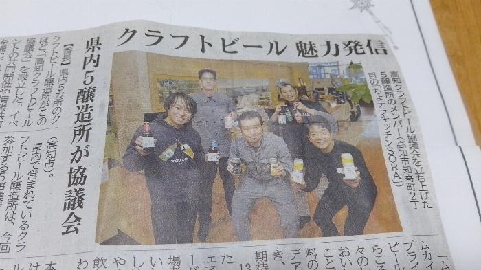 snippet from a Japanese newspaper featuring a photo of five brewers, all holding cans or bottles of their beers in a restaurant setting.